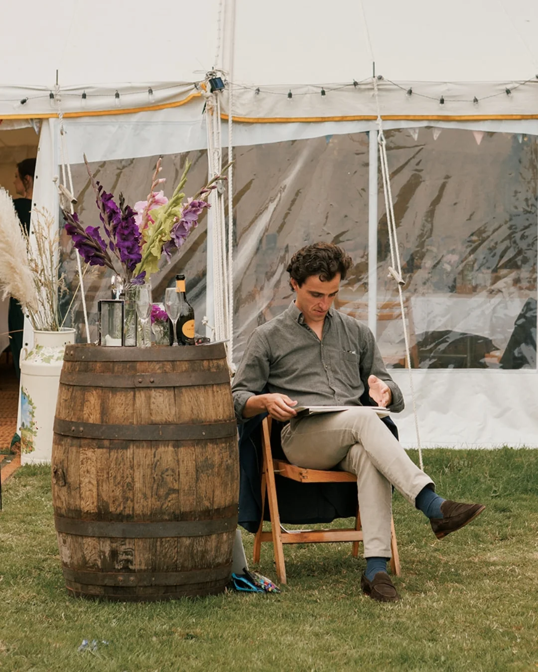 Rui Charnock seated on a folding chair at the edge of a marquee, head down, drawing on a board across his knee