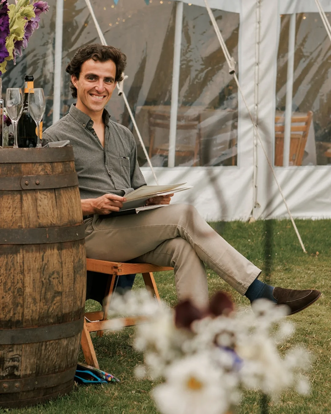 Rui Charnock seated on a folding stool beside a barrel inside a marquee at a wedding, an open sketchbook on his knee